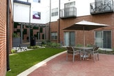 This outdoor amenity space is set within a well-designed courtyard enclosed by a multi-story residential building that combines classic red brick facades with contemporary white paneling and private balconies. The ground-level space is thoughtfully organized into distinct functional zones. A dedicated grilling station features a stainless steel propane grill mounted on a granite-topped counter, accompanied by posted instructions for communal use, emphasizing a community-oriented atmosphere. Nearby, a shaded seating area with tables, chairs, and large market umbrellas provides a comfortable outdoor dining and gathering spot. The courtyard features red-toned paver hardscaping complemented by lush plantings, ornamental shrubs, and mature trees that add greenery and natural character. A gated section leads to a swimming pool area, visible in the background alongside vibrant, colorful mural artwork on an adjacent parking structure. Smooth concrete walkways and black iron fencing define pathways and borders throughout the space. Large roll-up panel doors and low-profile windows at the building's base suggest access to additional interior amenity or utility spaces. The overall ambiance is communal, well-maintained, and designed for social engagement in an urban residential setting.