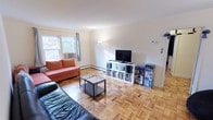 This single-floor space showcases a warm and functional living area defined by its classic parquet hardwood flooring, which lends a timeless character throughout. The living room is comfortably proportioned, offering ample space for seating and entertainment. A large window along one wall invites generous natural light, framing views of surrounding greenery and creating a bright, airy atmosphere.

The room accommodates a mix of seating, including an orange sofa and a dark leather couch, centered around a two-tier glass and metal coffee table. A white open-shelving media unit along the far wall serves as the entertainment and storage focal point, housing a flat-screen television, books, and various accessories. A floor lamp positioned nearby adds supplemental ambient lighting.

Baseboard heating runs along the lower walls, providing an efficient heating solution throughout the space. The white walls and neutral palette keep the interior feeling clean and open. The overall ambiance is casual and livable, with practical storage solutions and a straightforward layout that maximizes the usability of the space.