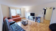 This single-floor space showcases a warm and functional living area defined by its classic parquet hardwood flooring, which lends a timeless character throughout. The living room is comfortably proportioned, offering ample space for seating and entertainment. A large window along one wall invites generous natural light, framing views of surrounding greenery and creating a bright, airy atmosphere.

The room accommodates a mix of seating, including an orange sofa and a dark leather couch, centered around a two-tier glass and metal coffee table. A white open-shelving media unit along the far wall serves as the entertainment and storage focal point, housing a flat-screen television, books, and various accessories. A floor lamp positioned nearby adds supplemental ambient lighting.

Baseboard heating runs along the lower walls, providing an efficient heating solution throughout the space. The white walls and neutral palette keep the interior feeling clean and open. The overall ambiance is casual and livable, with practical storage solutions and a straightforward layout that maximizes the usability of the space.
