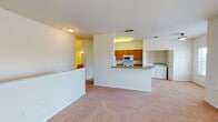This residence is accessed via a carpeted staircase with white-painted walls, leading up to the main living level. The upper floor opens into a spacious, open-plan living area finished with neutral wall-to-wall carpeting and textured ceilings. Large windows allow natural light to fill the space, creating a bright and welcoming ambiance throughout.

The kitchen is designed in a galley layout, featuring warm honey-toned wood cabinetry lining both sides, laminate countertops, a double stainless steel sink, and tile flooring. White appliances — including an electric range, microwave, and refrigerator — are fully integrated. A peninsula counter separates the kitchen from the living area, offering a casual pass-through connection between the two spaces.

Conveniently positioned off the main living area is a dedicated in-unit laundry alcove, housing a side-by-side washer and dryer. The bathroom is well-appointed with tile flooring, a single-sink vanity with matching wood cabinetry and stone-look countertop, and a full tub-shower combination with tiled surround. The bedrooms are carpeted and bright, each featuring windows that introduce natural light.