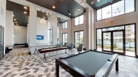Located at 3200 Peek Rd in Katy, TX, this single-story community clubhouse is a thoughtfully designed amenity space that blends contemporary style with practical functionality. The interior is defined by soaring double-height ceilings throughout, floor-to-ceiling windows that flood the space with natural light, and striking hexagonal tile flooring in a multi-tone palette that anchors the design aesthetic.

The game room is a standout feature, equipped with a full-size pool table and a shuffleboard table set against a backdrop of large windows overlooking an outdoor pool area. Pendant lighting with an industrial flair adds warmth and personality overhead. Adjacent to the game area, a casual lounge features deep sectional sofas accented with teal throw pillows, a dramatic geometric chandelier installation, and a textured stone accent wall with integrated sconce lighting — creating an inviting social atmosphere.

A dedicated meeting and co-working area provides a long conference table with ample seating, dual wall-mounted displays, and comfortable lounge chairs, making it well-suited for both collaborative work and informal gatherings. The space also includes a kitchenette and restroom facilities accessible via bold dark-paneled doors. Glass double doors connect the interior directly to outdoor areas, enhancing the open, connected feel of this well-appointed community hub.