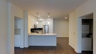 This single-floor space showcases a thoughtfully designed open-concept layout that seamlessly integrates the kitchen and living areas. The kitchen is a standout feature, equipped with sleek white cabinetry that extends to the upper walls, providing ample storage. Stainless steel appliances, including a full-size refrigerator and microwave, lend a modern, professional quality to the space. A gas range with a dark tile backsplash adds a bold contrast to the otherwise bright, neutral palette.

The kitchen island serves as a functional centerpiece, featuring a built-in sink and generous counter space finished in a clean white surface, ideal for both meal preparation and casual dining. Two elegant pendant lights hang above the island, adding focused illumination and a touch of sophistication.

Recessed lighting throughout ensures the space remains bright and well-lit. The dark hardwood-style flooring grounds the open layout and creates a warm contrast against the white walls and cabinetry. The adjacent living area offers a versatile, open canvas, completing this contemporary and practical space.