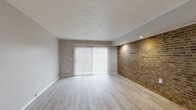 Located at 6847 Greenleaf Dr in Reynoldsburg, Ohio, this multi-level home spans three floors and offers a well-designed layout with 2 bedrooms, 3 bathrooms, and dedicated closet and utility spaces. The main living area features light hardwood-style flooring, a textured ceiling, and a striking full-length exposed brick accent wall illuminated by recessed lighting. A large sliding glass door with vertical blinds fills the space with natural light and offers outdoor views, creating an open and airy ambiance. The kitchen is outfitted with dark cabinetry, black appliances including a range and refrigerator, and a white quartz-style countertop, all complemented by the light flooring that carries throughout. Each of the three bathrooms showcases a consistent modern aesthetic with dark-framed vanities, white countertops, round mirrors, matte black fixtures, and light tile-style flooring. The staircase connecting the floors features warm wood treads, black metal railings, and carpet runners, adding a refined touch to the vertical circulation. The bedrooms are carpeted in a neutral tone and offer a calm, comfortable retreat. With three hallways connecting the floors and rooms, the layout ensures easy flow throughout the home.