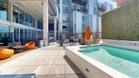 Located in the vibrant Shaw neighborhood of Washington, DC, this rooftop amenity level offers a thoughtfully designed outdoor retreat within an urban setting. The space is anchored by a pristine outdoor pool and adjacent spa, surrounded by wide-open tiled deck areas perfect for sunbathing, with lounge chairs and vibrant umbrella accents providing both shade and style. Lush green hedge walls add a natural contrast to the contemporary architecture, creating a sense of privacy within the city. The terrace transitions seamlessly into a covered outdoor lounge area framed by bold concrete columns and floor-to-ceiling glass walls, flooding the space with natural light and maintaining visual connectivity to the surrounding interiors. This lounge zone is furnished with sleek, modern sectional seating in cool blue tones arranged around a central fire pit table, ideal for gatherings during cooler evenings. Additional seating and dining areas are positioned throughout the terrace, offering a variety of settings for both casual and social use. The architectural design blends exposed concrete, metal paneling, and expansive glazing, giving the space an industrial-modern character. Surrounded by the dynamic Washington, DC skyline, this rooftop amenity space delivers a compelling combination of comfort, design, and outdoor living.