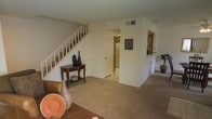 This two-story residence offers a functional and comfortable layout designed for everyday living. The main floor centers around a spacious carpeted living area that flows openly toward the kitchen, creating a welcoming and connected atmosphere. A white-railed staircase rises along one wall, providing a clean architectural transition to the upper level. The neutral-toned walls and light-colored carpeting throughout contribute to a bright and airy feel, while a textured ceiling adds subtle visual interest overhead. A pass-through or open wall section near the kitchen area allows for natural sightlines across the main living space, enhancing the sense of openness. The kitchen, visible from the living area, appears well-equipped and efficiently arranged. A dedicated wall niche near the entryway offers a practical display area, adding a decorative touch to the otherwise streamlined interior. HVAC vents are integrated into the ceiling and walls, ensuring climate comfort throughout. The overall design emphasizes a straightforward, livable floor plan with warm, neutral finishes that provide a versatile backdrop for a variety of furnishing styles. The second floor, accessible via the interior staircase, adds additional private living space above the main level.