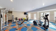 Located at 2615 Mt Zion Pkwy in Jonesboro, GA, this fitness center offers a thoughtfully designed exercise space across a single floor. The room is finished with warm taupe walls, white crown molding, and recessed lighting that creates a bright and welcoming atmosphere. A standout design feature is the bold, colorful hexagonal-patterned carpet in shades of blue, orange, and gray, which adds a dynamic and energetic feel to the space.

The fitness center is comprehensively equipped to support a variety of workout styles. Cardio equipment includes a treadmill and an elliptical machine, both positioned near large windows that allow natural light to fill the room and provide outdoor views during workouts. A stationary bike offers an additional cardio option. For strength training, the space features a multi-function cable machine, a dumbbell rack with a full range of weights, adjustable weight benches, and a selection of medicine balls and stability balls in various sizes. A wall-mounted flat-screen television is installed for entertainment during workouts.

The layout is open and well-organized, allowing ample room to move between stations. Access is provided through a white paneled door, and an exit sign is clearly visible, reflecting the space's attention to safety. Natural light streams in through multiple windows, enhancing the overall energy of this functional and inviting fitness facility.