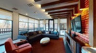 This well-appointed single-floor amenity space combines striking industrial design with modern functionality across several distinct areas. The centerpiece is a spacious lounge featuring exposed dark wood ceiling beams, dramatic accent lighting, and a bold brick-clad feature wall housing a wall-mounted TV and fireplace. Deep sectional sofas in charcoal gray, eclectic mixed seating, and unique stone-topped metal coffee tables create an inviting, social atmosphere. Floor-to-ceiling windows and glass doors wrap around the lounge, flooding the space with natural light and offering direct access to an outdoor terrace with views of surrounding trees. A business center adjacent to the lounge provides computer workstations with dedicated monitors, pendant lighting, and a mix of collaborative round tables with upholstered seating — all framed by large windows overlooking the terrace. The kitchenette is finished with sleek light-wood cabinetry, marble-look countertops, a subway tile backsplash, a sink, microwave, and under-counter refrigeration, accented by geometric pendant lights. A smaller meeting nook, decorated with framed Colorado-themed artwork, offers an intimate setting with terrace-facing windows and a direct door to the outdoor space, making this floor an ideal hub for both relaxation and productivity.