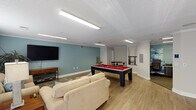 Located at 25 Landis Ln in Lillington, NC, this single-floor community amenity space is thoughtfully designed to offer residents a comfortable and engaging shared environment. The layout flows openly between several distinct functional zones, all united by light hardwood-style flooring, crown molding, and a neutral color palette accented by soothing teal walls.

The central game room is a standout feature, anchored by a full-size pool table with a bold red felt surface and a dedicated cue rack mounted on the wall. Nearby, a bistro-style high-top table with industrial-style stools provides a casual spot for socializing or spectating. A wall-mounted water fountain adds everyday convenience to this area.

Adjacent to the game room, a comfortable lounge area invites relaxation with a pair of plush, neutral-toned sofas accented with teal throw pillows, arranged around a patterned area rug. A large flat-screen television is mounted on the teal feature wall, and a decorative media console sits beneath it, completing the cozy setup. Natural light filters in through a windowed entry with glass-panel doors.

A dedicated workstation nook with a desk and ergonomic seating offers a quiet space for productivity. A teal built-in bookshelf unit nearby adds both storage and a decorative touch. The space is bright, well-lit with recessed fluorescent fixtures, and finished with textured ceilings and clean white trim throughout.