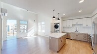 This thoughtfully designed single-floor residence offers an efficient and contemporary living experience. The open-concept layout seamlessly connects the living area and kitchen, creating a bright, airy atmosphere enhanced by large windows that invite ample natural light and views of the surrounding neighborhood.

The kitchen is a standout feature, showcasing a two-toned design with crisp white upper cabinetry and warm wood-textured lower cabinets, all complemented by a classic white subway tile backsplash. A central island with a clean white quartz countertop provides additional workspace and casual seating. Stainless steel appliances — including a refrigerator, range, microwave, and dishwasher — complete this functional and stylish space. Pendant lighting with matte black fixtures adds a modern touch overhead.

A convenient stacked washer and dryer unit is integrated within the main living area, adding practical in-unit laundry functionality. Light wood-tone flooring runs throughout, tying the spaces together cohesively.

The bedroom is a comfortable, well-proportioned retreat with a large window offering outdoor views and generous natural light. An adjacent walk-in closet features wire shelving along the walls, providing organized storage space. The overall palette of soft whites and neutral tones gives the unit a clean, modern, and welcoming ambiance.
