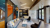 The lobby of RendezVous Urban Flats is a thoughtfully designed, single-floor communal space that blends modern urban aesthetics with functional amenities. Upon entry, floor-to-ceiling glass walls and black-framed glass partitions flood the space with natural light while providing clear sightlines to the surrounding streetscape. The reception area is anchored by a striking black feature wall bearing the RendezVous Urban Flats logo in bold gold lettering, paired with a sleek white reception desk that serves as the central point of welcome.

The lounge area is furnished with a generous sectional sofa accented by yellow patterned pillows, contemporary swivel chairs in charcoal tones, a low white coffee table, and soft round ottomans, all arranged atop a gray area rug over polished concrete floors. Linear LED lighting fixtures suspended from the ceiling provide a warm, modern glow throughout.

Adjacent to the lounge, a communal kitchen bar features a long stone-topped counter with decorative geometric tile detailing and bar stool seating, backed by large windows overlooking an outdoor area. A dedicated fitness zone with glass enclosure is also accessible from the main lobby. Exposed masonry walls with teal accent paintwork and framed artwork add character and visual interest to the overall design.
