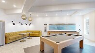 This thoughtfully designed amenity space offers residents a full suite of social and recreational areas across a single, well-appointed floor. The entrance lobby sets an inviting tone with geometric-patterned tile flooring, curated abstract artwork, lush potted greenery, and floor-to-ceiling windows that draw in abundant natural light and views of the streetscape beyond.

Adjacent to the entry, a spacious lounge area features soft, rounded blush-toned seating arranged around a sculptural marble coffee table atop a rich burgundy area rug. Large windows line the walls, filling the room with light, while carefully selected artwork and tall indoor plants add warmth and texture to the crisp white walls.

A sleek linear fireplace anchors a secondary lounge, complemented by gray upholstered armchairs, a bold yellow bench, and a mounted display screen — creating a cozy yet modern gathering space.

The community kitchen and dining area is outfitted with light wood cabinetry, open shelving, a full complement of appliances, and a central dining table, all illuminated by stylish pendant lighting.

The game room is a standout highlight, housing two light oak pool tables on dark tile flooring, a tufted yellow Chesterfield sofa, bistro seating, and a wall-mounted triptych of coastal artwork, all lit by ornate globe pendant fixtures that lend a warm, sophisticated ambiance.
