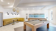 This thoughtfully designed amenity space offers residents a full suite of social and recreational areas across a single, well-appointed floor. The entrance lobby sets an inviting tone with geometric-patterned tile flooring, curated abstract artwork, lush potted greenery, and floor-to-ceiling windows that draw in abundant natural light and views of the streetscape beyond.

Adjacent to the entry, a spacious lounge area features soft, rounded blush-toned seating arranged around a sculptural marble coffee table atop a rich burgundy area rug. Large windows line the walls, filling the room with light, while carefully selected artwork and tall indoor plants add warmth and texture to the crisp white walls.

A sleek linear fireplace anchors a secondary lounge, complemented by gray upholstered armchairs, a bold yellow bench, and a mounted display screen — creating a cozy yet modern gathering space.

The community kitchen and dining area is outfitted with light wood cabinetry, open shelving, a full complement of appliances, and a central dining table, all illuminated by stylish pendant lighting.

The game room is a standout highlight, housing two light oak pool tables on dark tile flooring, a tufted yellow Chesterfield sofa, bistro seating, and a wall-mounted triptych of coastal artwork, all lit by ornate globe pendant fixtures that lend a warm, sophisticated ambiance.