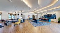 This generously sized amenity and recreation lounge in Huntsville, Alabama, offers a thoughtfully designed single-floor layout built for both leisure and social gatherings. The open-concept interior is finished with warm wood-look flooring throughout, crisp white walls, and a striking coffered ceiling with integrated ambient lighting that creates a polished, contemporary atmosphere.

The main lounge area centers around a billiards table set atop a bold blue area rug, surrounded by comfortable seating including a sectional sofa and accent chairs in vibrant yellow and blue tones. Stylish spherical pendant lights hang overhead, adding a modern artistic touch. Floor-to-ceiling glass doors lead to an outdoor balcony, flooding the space with natural light and offering views of the surrounding Huntsville community.

A dedicated Full Swing golf simulator room provides an immersive, high-tech experience complete with a turf hitting mat and large projection screen. Along the perimeter, a built-in window bench with rich burgundy upholstery and bistro-height tables offers a casual seating nook bathed in natural light.

The fully equipped kitchen features white cabinetry, a stainless steel refrigerator, a bold blue herringbone tile backsplash, and dark stone countertops — ideal for entertaining. A bar-height dining counter with colorful seating rounds out the social dining area, making this an all-inclusive space for recreation and community.
