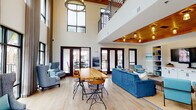 Located at 1813 Tyler Dr in Wilmington, NC, this two-story community clubhouse delivers a sophisticated blend of modern design and coastal-inspired décor. The main level greets visitors with soaring double-height ceilings, light hardwood flooring, and an abundance of natural light streaming through expansive floor-to-ceiling windows and French doors that open to an outdoor patio. A dramatic open-plan living area anchors the first floor, featuring a solid wood communal table, plush navy blue seating, and a warm wood tray ceiling with Edison-style pendant lighting. A dedicated lounge nook near the entrance offers a pair of wingback chairs ideal for quiet conversation. A full kitchen on the first floor is outfitted with white cabinetry, quartz countertops, a herringbone tile backsplash, globe pendant lights, wood ceiling beams, and a striking watercolor-style mural accent wall. An adjacent game room includes a full-size pool table, bar-height seating, and large windows. A sleek metal-railed staircase leads to the upper level, where exposed wood beams frame a bright secondary lounge and dining area complete with a gas cooktop island, gold-accented bar stools, and open views to the floor below.