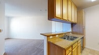 This single-story unit offers a practical and well-organized layout designed for comfortable everyday living. The galley-style kitchen is outfitted with honey-toned wood cabinetry, granite-style countertops, a double stainless steel sink, and black appliances including a refrigerator, electric range, and built-in microwave. A pass-through counter with an overhead wood panel visually connects the kitchen to the main living area, providing a casual bar seating option. The living space is finished with neutral wall-to-wall carpeting, creating a warm and cohesive feel throughout. A dedicated in-unit laundry closet houses a front-loading washer and accommodates a dryer, adding everyday convenience. The bedrooms are carpeted and receive natural light through large windows that offer views of the surrounding neighborhood. Wall-mounted heating units provide individual climate control. The bathroom features warm-toned tile flooring, a wood-framed vanity with ample storage, a large mirror with overhead lighting, and a full soaking tub with chrome grab bars and a shower curtain rod. The overall design is functional and understated, with consistent warm wood tones and neutral finishes throughout.