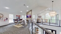 This single-floor community clubhouse is designed around a distinct aviation theme, combining modern comfort with bold, character-driven décor throughout. The centerpiece of the space is a spacious lounge area with soaring vaulted ceilings, abundant natural light from large multi-pane windows, and a sleek fireplace framed by a floor-to-ceiling accent wall. A wall-mounted flat-screen television anchors the room, while retro aviation artwork — including a prominent "National Air Races" poster — adds a spirited, curated personality to the space. Stylish egg-style chairs and a modern coffee table create an inviting conversation area, all set atop patterned carpet that adds texture and warmth. A cable-railed mezzanine-style overlook adds architectural depth and connects the lounge to an adjacent casual seating area filled with natural light from an arched window. The business center offers a long shared workstation with multiple computer terminals, a round café table, and ample natural light — ideal for productivity. The kitchen is fully equipped with dark cabinetry, granite countertops, black appliances including a full-size refrigerator and range, a peninsula with bar seating, pendant lighting, and aviation-themed artwork, providing a fully functional gathering and refreshment space for residents.