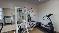 This single-floor fitness room offers a focused and functional workout environment designed to meet a variety of exercise needs. The space is outfitted with a Paramount multi-station cable and weight machine as its centerpiece, providing users with a wide range of strength training options including cable pulls, press stations, and resistance exercises. Complementing the strength equipment is a Precor elliptical trainer, offering low-impact cardiovascular exercise. Large wall-mounted mirrors line one side of the room, enhancing the sense of space while allowing users to monitor their form during workouts. The neutral-toned walls and carpeted flooring contribute to a clean, functional aesthetic. Overhead lighting ensures the space is well-lit throughout. The layout is efficiently organized to maximize usability within the available footprint, making it a practical amenity for those seeking convenient access to both strength and cardio equipment without the need for a full commercial gym.