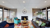 This single-floor space offers a warm and inviting atmosphere anchored by a standout living area. The room is centered around a classic fireplace set against a white brick accent wall, creating a charming focal point that adds character and coziness to the space. A large, decorative Roman numeral clock mounted above the fireplace adds a bold architectural detail that draws the eye upward to the drop ceiling fitted with recessed lighting, which provides a clean, evenly distributed illumination throughout the room. The flooring is finished in warm hardwood tones, lending a natural richness to the interior. Furnishings are vibrant and eclectic — a teal armchair, a deep brown leather sofa adorned with colorful throw pillows, and a floral-patterned area rug inject energy and personality into the space. A wooden coffee table with a decorative bowl serves as the centerpiece of the seating arrangement. Colorful abstract artwork adorns the walls, complementing the lively color palette. A dining area is also visible in the background, furnished with simple chairs and a table, making the space versatile for both casual gatherings and everyday use. Overall, the space balances comfort and character in a well-appointed, single-level layout.