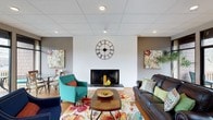 This single-floor space offers a warm and inviting atmosphere anchored by a standout living area. The room is centered around a classic fireplace set against a white brick accent wall, creating a charming focal point that adds character and coziness to the space. A large, decorative Roman numeral clock mounted above the fireplace adds a bold architectural detail that draws the eye upward to the drop ceiling fitted with recessed lighting, which provides a clean, evenly distributed illumination throughout the room. The flooring is finished in warm hardwood tones, lending a natural richness to the interior. Furnishings are vibrant and eclectic — a teal armchair, a deep brown leather sofa adorned with colorful throw pillows, and a floral-patterned area rug inject energy and personality into the space. A wooden coffee table with a decorative bowl serves as the centerpiece of the seating arrangement. Colorful abstract artwork adorns the walls, complementing the lively color palette. A dining area is also visible in the background, furnished with simple chairs and a table, making the space versatile for both casual gatherings and everyday use. Overall, the space balances comfort and character in a well-appointed, single-level layout.
