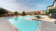 Located in Flower Mound, Texas, this outdoor community amenity space is designed to deliver a resort-like experience for its residents. At the heart of the space is a generously sized swimming pool with crystal-clear turquoise water, surrounded by a wide, open sundeck that accommodates rows of cushioned chaise lounges — perfect for relaxing under the Texas sun. The pool area is framed by mature trees and vibrant landscaping, adding a natural, welcoming atmosphere to the setting.

The surrounding courtyard is enclosed by an elegant wrought iron fence and anchored by multi-story Mediterranean-inspired buildings featuring warm stucco and stone facades, arched entryways, and private balconies. A dedicated patio lounge area features wicker seating grouped around tables with bold terracotta-colored umbrellas, providing ample shade for outdoor dining or socializing. A fire pit lounge area further enhances the outdoor experience, offering a cozy gathering spot. A covered veranda with decorative columns and a chandelier-style light fixture provides an additional sheltered seating area overlooking the pool. The overall ambiance is upscale, social, and tailored for outdoor living throughout the seasons.