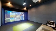 Located at 208 Windfall Way in Myrtle Beach, SC, this purpose-built indoor golf simulator room offers a fully immersive sports entertainment experience in a sleek, modern setting. The space is anchored by a large-format Full Swing Golf projection screen that spans the far wall, delivering high-definition, lifelike visuals of world-class courses and practice modes including driving range, target range, and short game options. A ceiling-mounted projector and track lighting illuminate the hitting area, which features a distinctive green artificial turf mat set against a dark, textured carpet floor — providing a clear and functional striking zone. The walls are clad in neutral, sound-absorbing panel materials that enhance the focused atmosphere of the room. A floor-to-ceiling dark curtain flanks the screen wall, lending a theater-like quality to the space. The lounge area transitions seamlessly from the simulator bay, furnished with a tufted tan leather sofa, cushioned ottomans, a small media console with a display screen, and a wall-mounted mini-split system for climate control. Dark painted walls and recessed ceiling lighting create a dim, sophisticated ambiance throughout. A glazed entry door provides access to the exterior, completing this well-appointed, single-floor simulation suite.