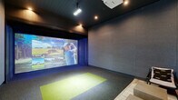 Located at 208 Windfall Way in Myrtle Beach, SC, this purpose-built indoor golf simulator room offers a fully immersive sports entertainment experience in a sleek, modern setting. The space is anchored by a large-format Full Swing Golf projection screen that spans the far wall, delivering high-definition, lifelike visuals of world-class courses and practice modes including driving range, target range, and short game options. A ceiling-mounted projector and track lighting illuminate the hitting area, which features a distinctive green artificial turf mat set against a dark, textured carpet floor — providing a clear and functional striking zone. The walls are clad in neutral, sound-absorbing panel materials that enhance the focused atmosphere of the room. A floor-to-ceiling dark curtain flanks the screen wall, lending a theater-like quality to the space. The lounge area transitions seamlessly from the simulator bay, furnished with a tufted tan leather sofa, cushioned ottomans, a small media console with a display screen, and a wall-mounted mini-split system for climate control. Dark painted walls and recessed ceiling lighting create a dim, sophisticated ambiance throughout. A glazed entry door provides access to the exterior, completing this well-appointed, single-floor simulation suite.
