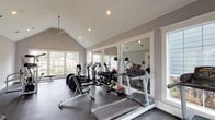 This fitness center is housed in a single-story space distinguished by soaring vaulted ceilings that lend an open, airy atmosphere throughout. The architectural design incorporates a prominent ridgeline ceiling accented by a central pendant ceiling fan, recessed lighting, and clean white trim work that contrasts neatly against the soft gray walls. Large multi-pane windows line multiple walls, flooding the interior with generous natural light and providing views of the surrounding greenery outdoors.

The flooring consists of durable dark rubber matting, well-suited for the demands of a fitness environment. A full wall of framed mirrors along one side visually expands the space while allowing users to monitor their form during workouts. The equipment selection is comprehensive, including multiple treadmills, elliptical cross-trainers, a rowing machine, and a stationary cycle for cardiovascular training. Strength-training options include a cable pulley machine, a multi-press station, weight benches, and a full rack of dumbbells ranging across various weight increments. Glass-paneled doors provide access to an adjoining area. The overall ambiance is clean, bright, and functional, offering a well-rounded environment suitable for a wide variety of fitness routines.