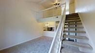 This two-floor residence is defined by its striking vertical openness, anchored by a soaring vaulted ceiling that rises above the main living area. A carpeted staircase with white-painted wooden railings leads up to an open loft level, which overlooks the living space below and is equipped with a ceiling fan for comfort. The loft railing mirrors the clean white aesthetic carried throughout the home, lending a cohesive and bright character to the interior.

The main living area is finished with wall-to-wall carpet in a neutral tone, creating a warm and inviting atmosphere. Natural light filters into the space, enhancing the sense of openness created by the double-height ceiling. A kitchen is situated at the rear of the main floor, partially open to the living area via a pass-through counter. It features dark appliances, including a full-sized refrigerator, and is illuminated by a chandelier-style light fixture that adds a touch of classic charm.

The overall design balances practicality with an open, airy feel. The loft configuration provides flexible use of the upper level, whether as a sleeping area, home office, or additional living space. The consistent use of white trim, neutral carpeting, and warm lighting gives the home a clean and welcoming ambiance throughout both floors.