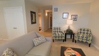 This single-floor residence offers a thoughtfully arranged living space with a warm, comfortable ambiance. The living area serves as the central hub of the home, featuring soft wall-to-wall carpeting in a neutral beige tone that complements the light gray walls throughout. A plush sofa anchors the seating area, accented by a patterned accent chair in bold navy and green geometric print, adding a touch of visual interest to the otherwise understated palette. A dark wood side table topped with a classic table lamp and small greenery provides both function and warmth to the space.

Wall décor includes a framed mirror with a dark border and two framed prints, lending a curated, finished feel to the room. A ceiling vent is positioned above, indicating a central HVAC system for climate control.

The open hallway beyond the living area connects to additional rooms, including what appears to be a bedroom with a visible closet and a bathroom, ensuring a practical flow throughout the unit. The front entry door is visible at the end of the corridor, reinforcing the straightforward, single-level layout. Overall, the space presents a clean, move-in-ready environment with a modest and welcoming character.