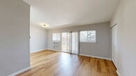 This single-floor unit offers a thoughtfully designed layout spread across a comfortable footprint. The open-concept living area is finished with warm, light-toned hardwood flooring that extends throughout the main living spaces, creating a cohesive and inviting atmosphere. Large sliding glass doors lead to a private balcony, flooding the living room with natural light and providing outdoor views, while an additional window further brightens the space.

The kitchen is neatly defined within its own alcove and features white shaker-style cabinetry, granite-look countertops, a stainless steel sink, and black appliances including a full-size refrigerator and gas range. A skylight above adds an extra source of natural light to the cooking area.

The bedroom is carpeted for comfort and includes a built-in closet with bifold doors, as well as a window that allows for natural light and outdoor views. Additional rooms throughout the unit are also carpeted and offer versatile usage potential, each featuring neutral gray wall tones, white trim, and ample natural light from multiple windows.

Wall-unit air conditioners are present in several rooms, and clean white doors and trim provide a crisp, modern finish throughout the entire space.