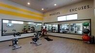 This single-floor fitness facility is designed to energize and inspire members from the moment they walk in. The workout area features a clean, open layout with light wood-style flooring that enhances the bright, welcoming atmosphere. Large wall-to-wall mirrors line the perimeter, visually expanding the space and allowing members to monitor their form during exercise. High ceilings with recessed lighting ensure the area is well-lit, while elevated windows allow natural light to filter in, adding to the airy feel. Bold yellow accent stripes contrast against crisp white walls, reinforcing the facility's vibrant and motivating aesthetic. A standout wall phrase — "Make Muscles. Not Ex..." — sets an encouraging tone throughout the space. The gym is outfitted with cycling stations, medicine balls, and a variety of strength and cardio equipment, making it suitable for a wide range of fitness routines. A mounted flat-screen TV keeps members entertained during workouts. The thoughtful combination of functional equipment, reflective surfaces, and energetic design elements creates an environment well-suited for both individual training sessions and group fitness activities.