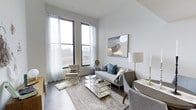 This single-floor living space is designed with a contemporary aesthetic, emphasizing light, openness, and refined style. The living area is anchored by a pair of large, dark-framed windows dressed with sheer white curtains that allow natural light to flood the room, illuminating the soft grey and white color palette throughout. The windows offer views of the surrounding urban landscape, reinforcing the space's connection to the outside environment.

The room features dark hardwood flooring that grounds the otherwise light interior, providing a subtle contrast to the pale walls. A plush area rug with a muted, traditional pattern defines the seating zone, layering texture and warmth into the space. Furnishings include a grey upholstered sofa accented with blue throw pillows, a cream accent chair, and a gold-framed glass coffee table that adds a touch of glamour. Complementary gold-toned table lamps and décor accessories reinforce a cohesive, modern-glam design language. A large abstract artwork in muted tones serves as a focal point on the main wall, tying the color scheme together and adding artistic character to this well-appointed living space.