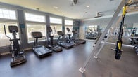 This single-floor fitness center offers a clean, functional environment designed with exercise and wellness in mind. The space is outfitted with a row of modern cardio machines, including treadmills and ellipticals, arranged to maximize floor space and provide an efficient workout layout. Durable, dark rubber flooring covers the exercise area, providing both safety and comfort underfoot. A large wall-mounted mirror spans one side of the room, creating a sense of openness while allowing users to monitor their form during workouts. The room is brightened by a series of large windows along one wall, inviting generous amounts of natural light and offering views of the surrounding outdoor greenery. Recessed ceiling lighting and ceiling fans supplement the natural light and help maintain a comfortable temperature throughout the space. The walls are painted in a soft gray-blue tone, accented by teal trim details, lending the room a fresh and motivating atmosphere. The overall design balances practicality with comfort, making this fitness center a well-considered amenity space suited for a range of cardio-focused exercise routines.