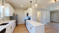 This well-appointed two-floor residence showcases a clean, contemporary design throughout. The heart of the home is a galley-style kitchen featuring crisp white cabinetry adorned with brushed gold hardware, striking quartz countertops with subtle veining, and a full suite of stainless steel appliances including a refrigerator, range, built-in microwave, and dishwasher. A central island provides additional prep and storage space, while pendant lights with glass jar shades hang above, adding a stylish finishing touch. Light wood-tone flooring runs throughout the main living areas, lending warmth and continuity to the open-concept layout. The living area is generously sized with a ceiling fan overhead, neutral walls, and natural light filtering in from nearby windows. The kitchen transitions seamlessly into this space, creating an ideal flow for everyday living. The bedrooms feature plush carpeting and ceiling fans, offering a comfortable and quiet retreat. The bathrooms are tastefully finished — one includes a walk-in shower with built-in shelving and wood-look flooring, while another offers a full tub-shower combination with a white vanity and gold-accented fixtures. Natural light brightens each bathroom through well-placed windows. Overall, the space presents a fresh, move-in-ready aesthetic with consistent modern finishes across both floors.