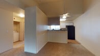 This single-floor residence offers an open and functional layout that combines the living and kitchen areas into one cohesive space. The vaulted ceilings add a sense of height and openness, while a ceiling fan with integrated lighting provides both comfort and illumination. The kitchen is defined by a wraparound peninsula featuring classic white beadboard paneling and a dark countertop, creating a clean contrast. White upper cabinetry lines the kitchen walls, complemented by a black refrigerator and a window above the counter that allows natural light to filter in. The kitchen transitions smoothly into the living area, which is finished with warm, neutral carpeting that extends throughout. A partial wall with matching beadboard detailing helps delineate the two spaces while maintaining the open feel of the floor plan. Hardwood-style flooring is visible near the kitchen, adding a practical and visually appealing touch to the cooking area. The overall ambiance is casual and welcoming, with a neutral color palette of soft whites and warm beiges that creates a bright, move-in-ready environment well-suited for comfortable everyday living.