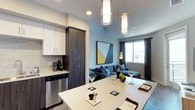 This single-floor residence showcases a thoughtfully designed open-concept layout that seamlessly connects the kitchen and living areas. The kitchen is a standout feature, equipped with stainless steel appliances including a full-size refrigerator, range, microwave, and dishwasher. Two-tone cabinetry pairs dark wood-grain lower cabinets with crisp white upper cabinets, complemented by a tiled backsplash and light quartz countertops. A central island with pendant lighting above serves as both a prep space and casual dining area.

The living area flows naturally from the kitchen, anchored by a comfortable sectional sofa and accented with bold geometric artwork. Natural light enters through large windows dressed with dark curtain panels, creating a balanced, modern ambiance with soft blue accent walls adding warmth.

The bedroom offers a serene retreat with plush carpeting, large windows, and a generously sized bed with layered navy and grey bedding. The bathroom features wood-look flooring, a double-sink vanity with dark cabinetry and white countertops, a large mirror, and a full soaking tub with shower. Together, these spaces present a cohesive, contemporary aesthetic throughout.