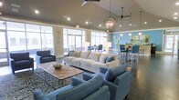 This single-floor space offers an open-concept layout designed for comfortable communal gathering. The lounge area is anchored by soaring vaulted ceilings that lend a sense of spaciousness and airiness throughout. Large floor-to-ceiling windows line the perimeter, flooding the interior with abundant natural light and providing views to the outdoors. The main seating area features a generously sized neutral-toned sectional sofa complemented by coordinating blue accent chairs arranged around a metal-framed coffee table, all set atop a patterned area rug over polished concrete flooring. Overhead, a combination of ceiling fans and a distinctive spherical pendant light fixture add both function and visual interest. Adjacent to the lounge, a bar-height dining area is set against a bold blue accent wall adorned with decorative wall art, furnished with high-top chairs in coordinating blue tones. Recessed lighting throughout ensures the space remains well-lit at all times. The overall ambiance is modern and inviting, with a cohesive blue and neutral color palette that ties together the various zones, making the space well-suited for socializing, casual meetings, or relaxed everyday use.