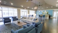 This single-floor space offers an open-concept layout designed for comfortable communal gathering. The lounge area is anchored by soaring vaulted ceilings that lend a sense of spaciousness and airiness throughout. Large floor-to-ceiling windows line the perimeter, flooding the interior with abundant natural light and providing views to the outdoors. The main seating area features a generously sized neutral-toned sectional sofa complemented by coordinating blue accent chairs arranged around a metal-framed coffee table, all set atop a patterned area rug over polished concrete flooring. Overhead, a combination of ceiling fans and a distinctive spherical pendant light fixture add both function and visual interest. Adjacent to the lounge, a bar-height dining area is set against a bold blue accent wall adorned with decorative wall art, furnished with high-top chairs in coordinating blue tones. Recessed lighting throughout ensures the space remains well-lit at all times. The overall ambiance is modern and inviting, with a cohesive blue and neutral color palette that ties together the various zones, making the space well-suited for socializing, casual meetings, or relaxed everyday use.
