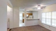 This single-story space features an open and functional layout that connects the living area to the kitchen through a wide pass-through counter, creating a sense of openness and easy interaction between spaces. The living area is finished with wall-to-wall carpet and includes a ceiling fan with integrated lighting, providing both comfort and airflow. A window along the side wall allows natural light to brighten the room.

The kitchen is accessible via a separate entryway and is visible from the living area through the pass-through opening. It features warm-toned wood cabinetry, white appliances including a refrigerator and microwave, and a light-colored countertop that extends to form the pass-through bar. The kitchen benefits from its own window, adding additional natural light to the cooking space. The flooring in the kitchen is a smooth, light-toned surface, contrasting with the carpeted living area.

Throughout the space, white walls and a textured ceiling contribute to a clean, neutral aesthetic. The layout is practical and straightforward, making efficient use of the available space while maintaining a connected, cohesive feel between the main living and kitchen areas.