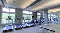 This fitness center is designed to provide a comprehensive workout environment within a single-level layout. The space is filled with an abundance of natural light streaming in through a row of large windows that line the upper walls, offering pleasant views of the surrounding greenery and creating an open, energizing atmosphere. The neutral-toned walls are accented by exposed ductwork along the ceiling, lending the room a modern, industrial aesthetic. A full-length mirror spans one wall, visually expanding the space and allowing users to monitor their form during exercise. The cardio section is well-appointed with multiple treadmills and elliptical machines, all positioned to take advantage of the outdoor views. A weight bench is also present, supporting strength training alongside the cardio equipment. The flooring is carpeted, providing comfort underfoot throughout the facility. The overall ambiance is clean, functional, and well-lit, making it a practical and motivating environment for a wide range of fitness activities.
