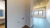 Located at 2350 Saratoga St in Alameda, California, this single-floor live/work unit showcases a compelling blend of industrial heritage and contemporary design. The space is defined by its dramatic vaulted ceilings with exposed wood beams and concrete structural elements, lending an authentic loft character throughout.

The open-plan living area features polished concrete floors and an arched ceiling detail supported by a prominent concrete column, creating a striking architectural focal point. Large, multi-pane industrial-style windows flood the space with abundant natural light while offering views of the surrounding Alameda neighborhood.

The kitchen is well-appointed with warm wood-tone cabinetry, light subway tile backsplash, and a full suite of stainless steel appliances including a refrigerator, wall oven, electric cooktop, built-in microwave, and dishwasher. A wall-mounted mini-split HVAC unit ensures efficient climate control throughout the space.

The bathroom features clean, modern finishes including white subway tile surrounding a soaking tub and shower combination with accessibility grab bars, a floating vanity with integrated sink, and stone-look tile flooring. An electrical panel and utility connections are conveniently accessible within the unit. The overall ambiance is urban and industrial, yet refined — ideal for creative or professional use.