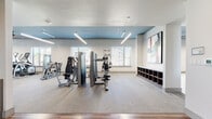 This fitness center is thoughtfully designed to accommodate a wide range of workout needs within a single-level layout. The space is divided into distinct functional zones, each tailored to a specific type of training. The main cardio area hosts a row of treadmills and elliptical machines aligned along windowed walls, allowing natural light to filter in through slatted blinds. A tray ceiling painted in a bold teal-blue adds visual definition to this zone, complemented by ceiling fans and linear pendant lighting that contribute to a clean, contemporary aesthetic.

Adjacent to the cardio section, the strength training area features multiple cable and weight machines arranged to maximize floor space, with large framed mirrors along the walls enhancing the sense of depth. Striped low-pile carpet covers much of the workout floor, providing cushioning and grip underfoot, while hardwood-style flooring transitions into the studio zone.

The dedicated studio room is notably open, finished with light hardwood-style flooring, a teal accent wall, and a wall-mounted display screen for guided workouts. A ceiling fan and linear light fixture complete the space. A separate area with pass-through mirrors provides views into the free weights section, which includes medicine ball storage and additional equipment, rounding out this comprehensive fitness facility.