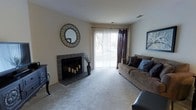 This single-floor residence offers a thoughtfully arranged layout with distinct living and kitchen zones. The living room is a comfortable, carpeted space anchored by a tile-surround fireplace, creating a natural focal point. A large sliding glass door with vertical blinds and drape panels floods the room with natural light while providing access to an outdoor area. The open sightlines from an adjacent passageway connect the living room to the rest of the unit seamlessly, and a framed art piece adds a decorative touch to the neutral-toned walls.

The galley-style kitchen is efficiently designed with white cabinetry lining both walls, offering ample storage. It features black appliances, including a range with an overhead exhaust hood and a full-size refrigerator, alongside a dishwasher and a double-basin sink. Dark tile flooring grounds the space. A doorway at the end of the kitchen leads to a dedicated laundry area, where a washer and dryer are conveniently housed within the unit.

Overall, the residence maintains a neutral color palette throughout, with light walls and carpeted floors in the living areas, creating a cohesive and understated ambiance.