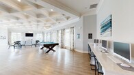 Located at 1001 Cox Grade Rd in Panama City Beach, FL, this single-story community clubhouse presents a refined coastal aesthetic throughout its thoughtfully designed spaces. Warm hardwood-style flooring flows seamlessly from room to room, complemented by crisp white trim, crown molding, and soft neutral wall tones that create a welcoming, airy atmosphere.

The main gathering area is a standout, featuring a striking coffered ceiling with geometric detailing and an elegant ring chandelier. Expansive windows dressed in botanical-print drapes flood the space with natural light and offer views of the surrounding community. Teal leather swivel chairs and plush seating arrangements invite relaxed conversation, while a wall-mounted television adds a modern entertainment element.

A dedicated game room centers around a full-size pool table beneath the same impressive coffered ceiling, with an extended bar-height counter and seating along one wall — ideal for social gatherings. Adjacent to this, a bright kitchen-style area features a long white quartz countertop island with pendant lighting above, bar seating, and coastal artwork on the walls.

A charming dining nook accommodates an ornate round table with elegantly carved chairs, surrounded by large windows that bathe the room in natural light. A wide, well-lit hallway with recessed lighting connects all areas, completing this polished and functional community space.