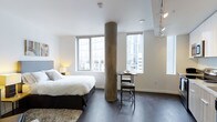 This single-floor studio is a thoughtfully designed open-plan space that blends modern aesthetics with urban living. The most distinctive architectural feature is a striking cylindrical concrete column positioned near the center of the room, serving as a natural visual divider between the sleeping and dining areas while adding an industrial character to the overall design.

Dark hardwood floors run throughout the space, providing a rich contrast against the crisp white walls and ceilings. The sleeping area accommodates a full-sized bed dressed in neutral linens, complemented by a warm bedside lamp that adds a cozy ambiance. A textured area rug anchors the sleeping zone, adding softness underfoot.

Adjacent to the column, a compact bistro-style table and stool seating arrangement creates a defined dining nook. Two generously sized windows allow abundant natural light to fill the space and frame views of the surrounding urban skyline, reinforcing the city-connected atmosphere.

Overhead, a track lighting fixture provides additional directional illumination, enhancing the studio's contemporary appeal. The overall ambiance is clean, minimal, and functional, making efficient use of the open layout.