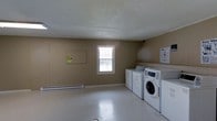 This single-floor laundry facility is a straightforward, utilitarian space designed to meet everyday laundry needs. The room features a row of commercial-grade washing machines and dryers lined along one wall, providing ample capacity for multiple users. The machines appear to be coin- or card-operated, as is typical of shared laundry facilities.

The interior is finished with neutral tan-painted walls and a textured popcorn ceiling, giving the space a simple, no-frills aesthetic. A single window with a white frame allows natural light to filter into the room, brightening the otherwise modest interior. Barred for security, the window also provides a degree of ventilation.

The flooring is a light-colored, easy-to-clean surface suited to the functional demands of a laundry room. A baseboard heating unit runs along the lower portion of one wall, ensuring the space remains comfortable during cooler months. Informational notices and posted signage are visible on the walls, providing guidance for users. Overall, the space is clean, practical, and efficiently organized to serve its intended purpose.