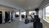 This fitness center is designed to support a well-rounded workout experience within a single-level layout. The space features a neutral color palette with light grey walls and carpeted flooring, creating a clean and functional atmosphere. Large windows along the perimeter allow natural light to fill the room, offering views of the surrounding outdoor area and contributing to an open, airy feel. Structural columns are integrated into the layout without significantly impeding the usable floor space. The cardio section is equipped with modern machines, including a treadmill, elliptical, and stair climber, positioned to take advantage of the natural light from the windows. A wall-mounted television is available for entertainment during workouts. The free weights area features multiple dumbbell racks stocked with a range of weights, accompanied by a flat workout bench for strength training exercises. Mirrored walls on one side of the room enhance the sense of space while allowing users to monitor their form. A wall clock is conveniently positioned for easy time-tracking. Recessed lighting supplements the natural light, ensuring the space remains well-lit throughout the day.