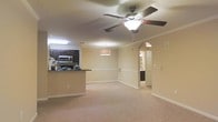 This single-floor residence offers a thoughtfully designed open-concept layout that connects the living area and kitchen in a seamless, airy arrangement. The spacious living area is finished with plush wall-to-wall carpeting and features a ceiling fan with an integrated light fixture, providing both comfort and functionality. Crown molding runs along the upper walls, lending a refined architectural touch throughout the space.

The kitchen is defined by rich, dark wood cabinetry that contrasts elegantly with the light-toned walls and countertops. A breakfast bar serves as a natural divider between the kitchen and living area, making it ideal for casual dining or entertaining. Stainless steel appliances, including a microwave, are integrated into the cabinetry for a cohesive look.

Warm, neutral tones dominate the interior palette, creating a welcoming and versatile ambiance. An arched entryway on one side of the living area adds a classic architectural detail while guiding the eye toward additional rooms beyond. The additional spaces glimpsed through the arch suggest further well-appointed areas, including what appears to be a hallway leading to additional rooms. Overall, the space balances practical design with tasteful finishes, making it a comfortable and functional living environment.