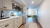 This single-floor studio is a smartly organized open-plan space that maximizes every square foot through clever zoning and contemporary design choices. The kitchen is a standout feature, outfitted with shaker-style cabinetry in a warm taupe finish, a crisp white countertop, a black undermount sink, stainless steel appliances including an over-the-range microwave, and a striking patterned tile backsplash that adds visual interest and character. Warm hardwood-style flooring flows seamlessly throughout the entire space, unifying each zone. A compact dining area accommodates a hairpin-leg table paired with teal accent chairs, creating a vibrant and inviting spot beneath tasteful wall art. The living area is anchored by a sliding barn door and a dark open-shelving unit that acts as a natural divider between the living and sleeping zones, providing both storage and a sense of separation without closing off the open feel. The sleeping area is comfortably appointed and benefits from natural light through a large window. The bathroom features a clean, modern aesthetic with hexagonal dark tile flooring, a white vanity with a vessel sink, matte black hardware and fixtures, and a subway-tiled shower, delivering a cohesive and polished finish throughout.