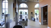 This single-story fitness center is designed to provide an open, energizing workout environment. The space is defined by its striking architectural character, most notably the tall, arched floor-to-ceiling windows that line the exterior walls, flooding the interior with natural light and offering views of the surrounding greenery and outdoor areas. The high ceilings contribute to a spacious, airy atmosphere that enhances the overall workout experience.

The facility is well-appointed with a range of cardio equipment, including elliptical trainers and a rowing machine, all positioned to take advantage of the natural light and outdoor views. The equipment is modern and features integrated display screens for interactive workout tracking. The neutral, light-toned walls and carpeted flooring create a clean, comfortable aesthetic throughout the space.

Subtle decorative wall panels add a touch of visual interest without distracting from the functional nature of the room. Exposed industrial-style ductwork along the ceiling reinforces the contemporary design sensibility. The layout is open and well-organized, allowing for easy movement between equipment and providing a practical, user-friendly workout space.